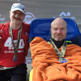 Two men pose after a race. One stands wearing a red shirt, medal, and hat, smiling with his arm around the other man, who sits in a wheelchair, covered in an orange blanket, also wearing a medal. A race banner is behind them.