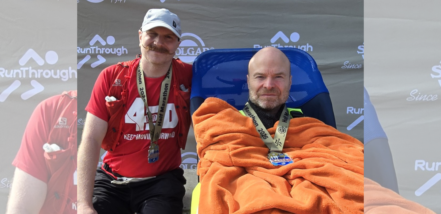 Two men pose after a race. One stands wearing a red shirt, medal, and hat, smiling with his arm around the other man, who sits in a wheelchair, covered in an orange blanket, also wearing a medal. A race banner is behind them.