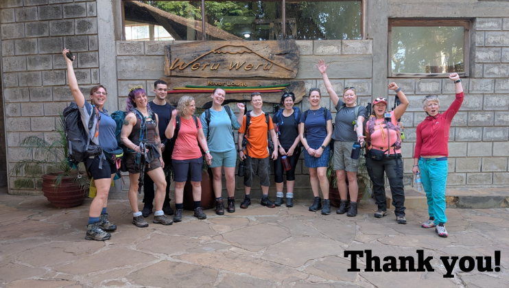 A group of hikers, wearing outdoor gear and backpacks, smile and raise their arms in celebration outside the Weru Weru River Lodge. 