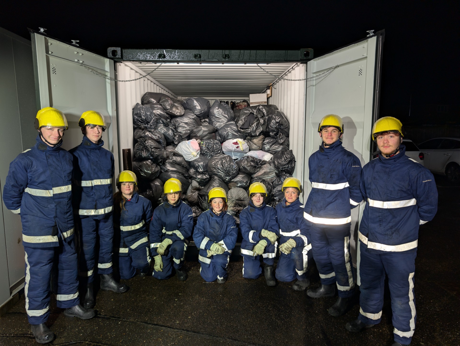 Nine people in blue uniforms and yellow helmets stand and kneel in front of an open shipping container filled with tightly packed black garbage bags at night.