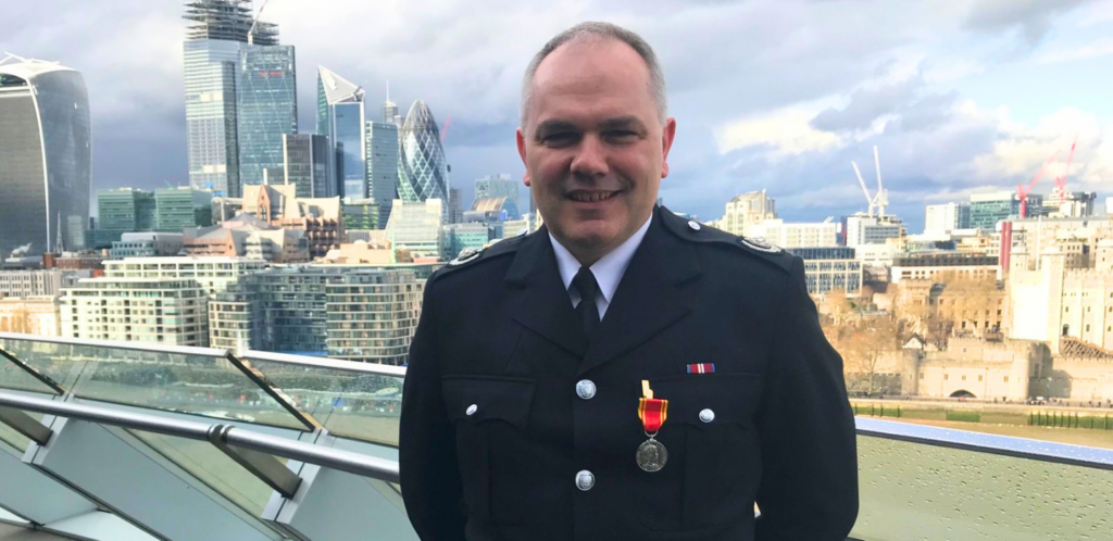 A portrait of Steve Norman, a former Deputy Assistant Commissioner for London Fire Brigade, smiling in an outdoor setting. in front of the London city skyline.