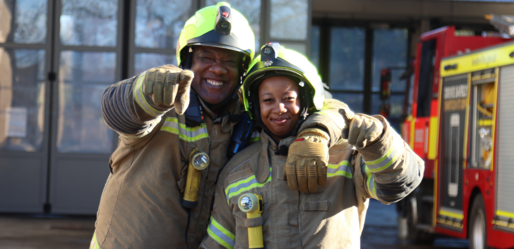 Two firefighters in protective gear smile and point toward the camera, standing close together in front of a fire truck and building.