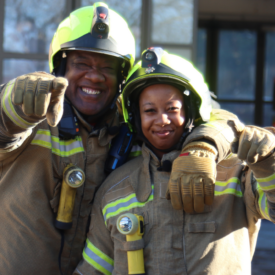 Two firefighters in protective gear smile and point toward the camera, standing close together in front of a fire truck and building.