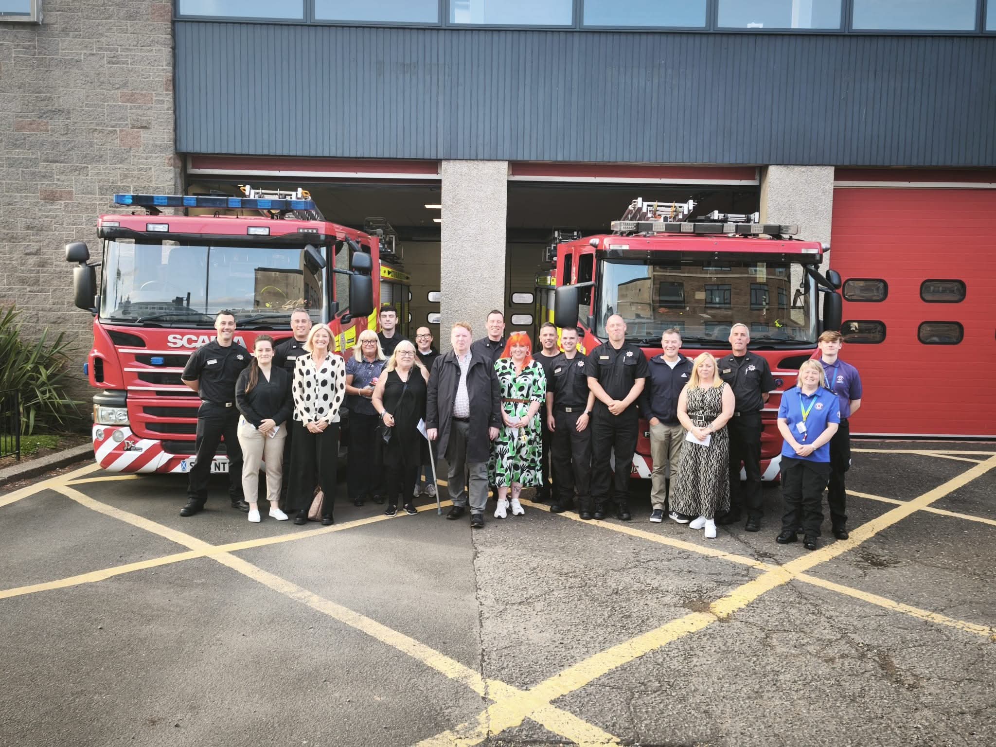 A group of people, including firefighters in uniform and others in casual clothing, stand smiling in front of two red fire trucks parked outside a fire station.