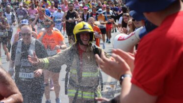 A firefighter in full gear smiles while running in a crowded marathon, as people on the sidelines cheer and spray water, creating a festive, spirited atmosphere.