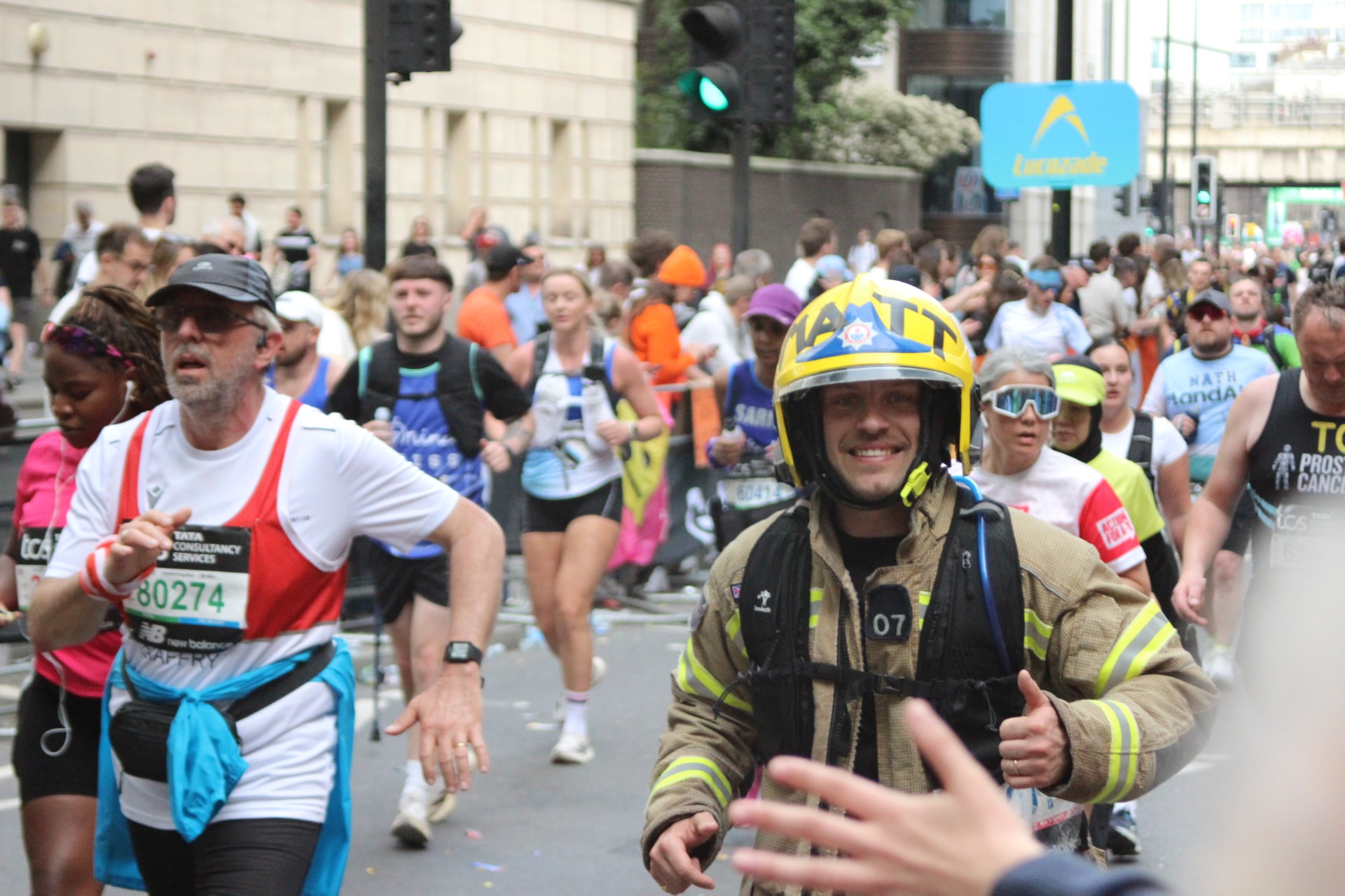 A crowd of marathon runners moves down a city street. One runner in the foreground wears a firefighter’s uniform and helmet, smiling and giving a thumbs-up. Other runners wear athletic gear and race bibs. Spectators line the route.
