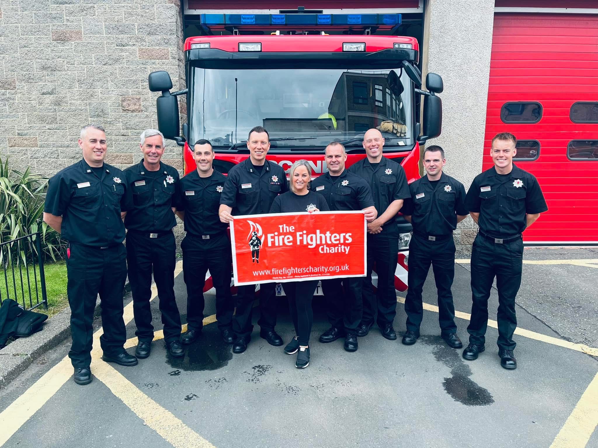 A group of eight firefighters in uniform and one woman stand in front of a fire truck, smiling and holding a red banner that reads "The Fire Fighters Charity." A red garage door is in the background.