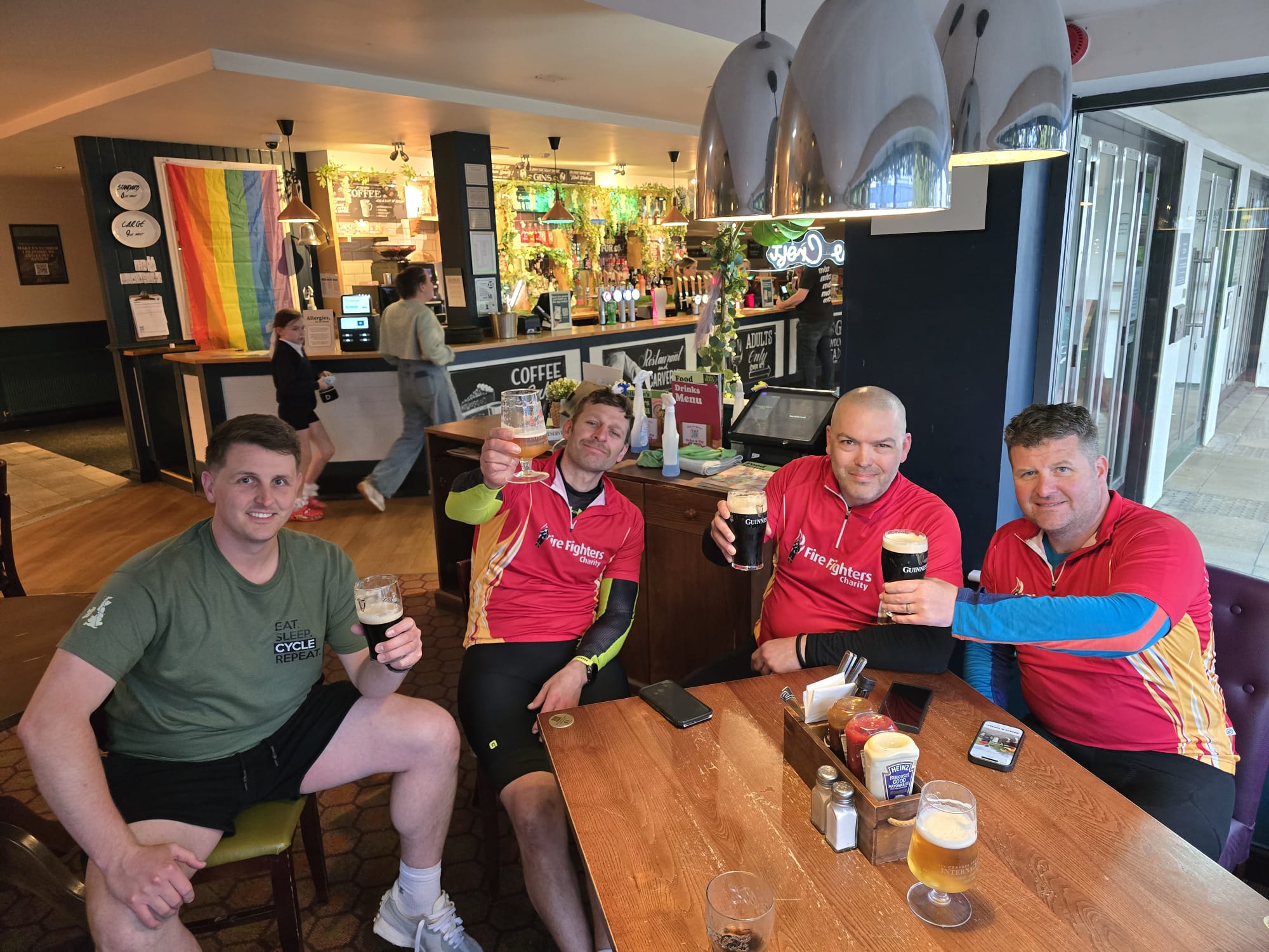 Four men in cycling gear are sitting around a pub table, smiling and raising pints of beer. Behind them is a bar with a rainbow flag, drinks, and decorations. One person is standing at the bar in the background.