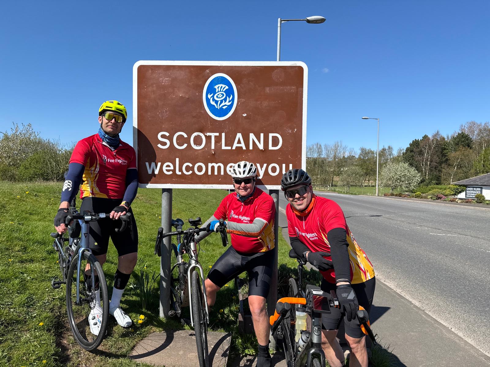 Three cyclists in red jerseys pose with their bikes in front of a large brown 
