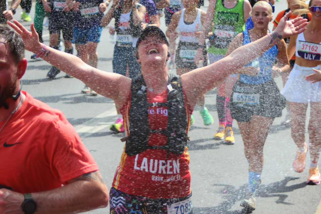 A marathon runner in a red tank top with "LAUREN" on it smiles joyfully with arms raised as water sprays over her. Other runners are visible in the background, all wearing race bibs and athletic clothing.