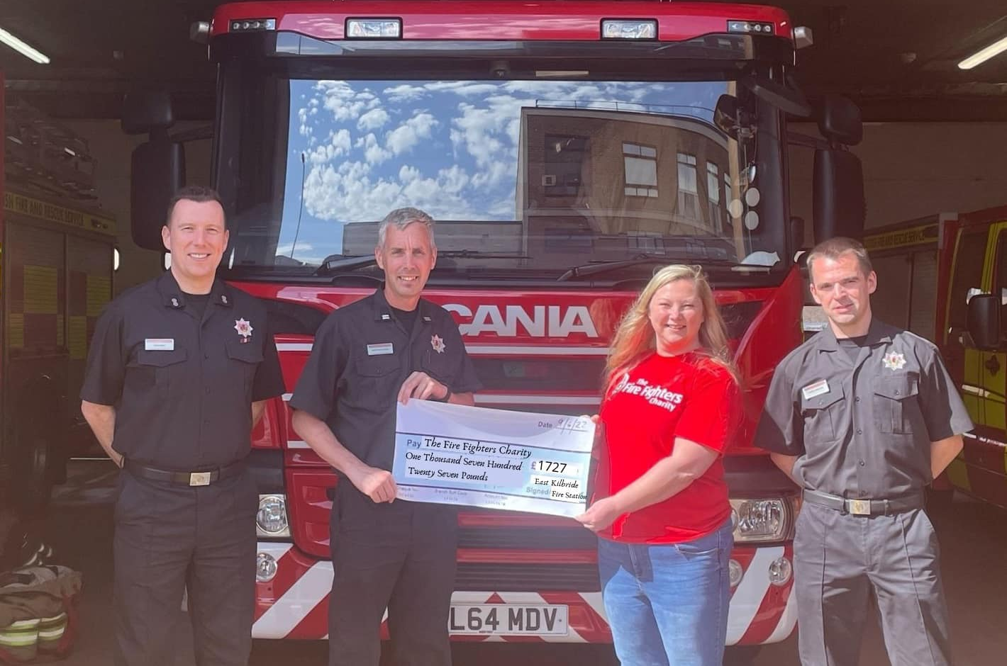 Four people stand in front of a fire engine; three wear dark fire service uniforms, and one in a red shirt holds a large donation cheque with them, smiling for the camera. Sunlight brightens the scene.
