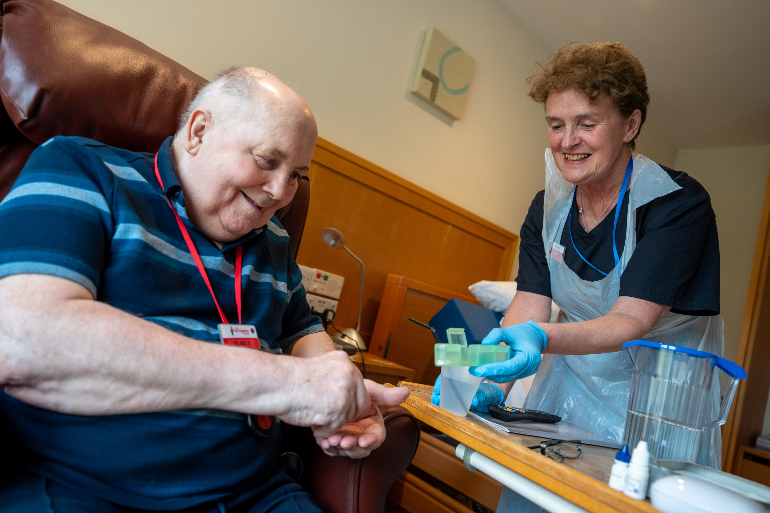 An elderly man sits in a chair smiling as a nurse in gloves and an apron prepares to take a blood sample from his finger in a well-lit room with medical supplies on a tray. 