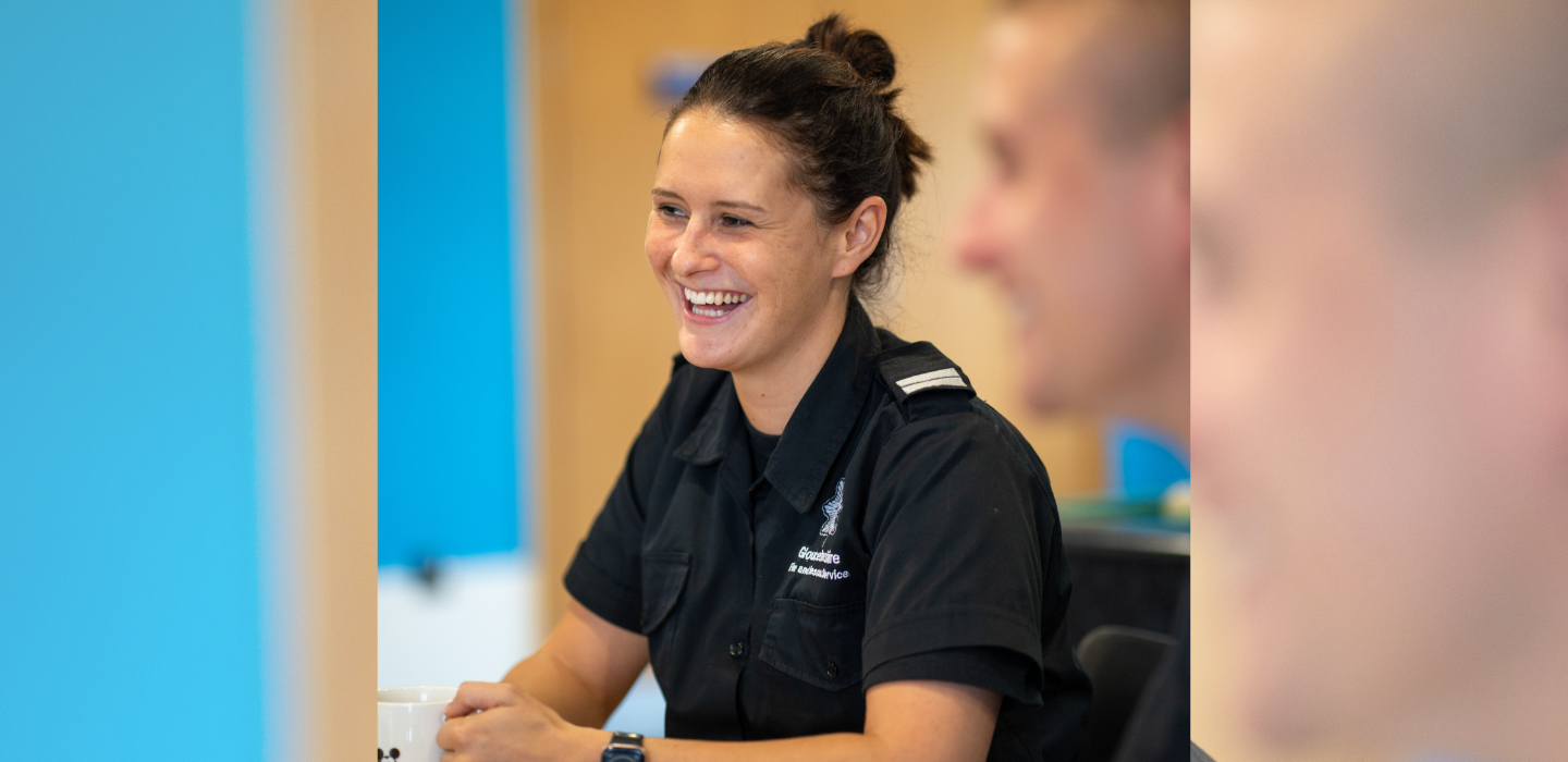 A smiling woman in a dark uniform sits at a table, holding a mug. The background is softly blurred, and another person is partially visible in the foreground.