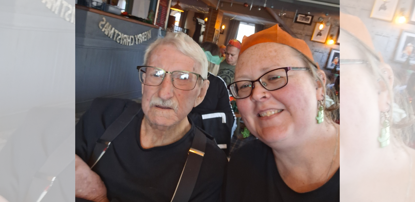 An older man with glasses and a woman wearing glasses and a paper crown smile for a selfie in a festive, decorated room with a 