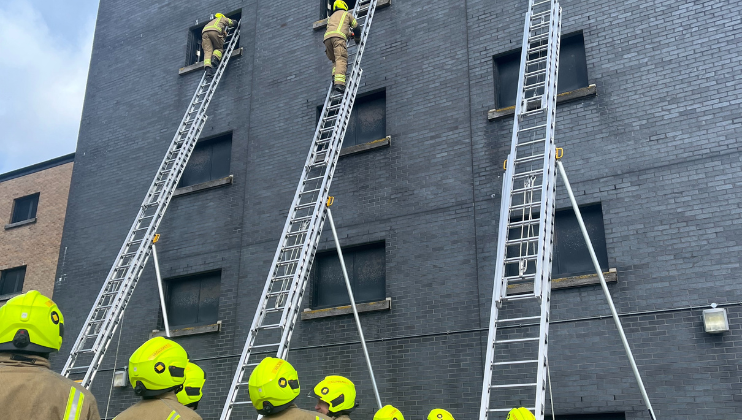 Firefighters wearing bright yellow helmets stand on the ground, watching as two firefighters climb tall ladders leaning against a grey brick building during a training exercise.