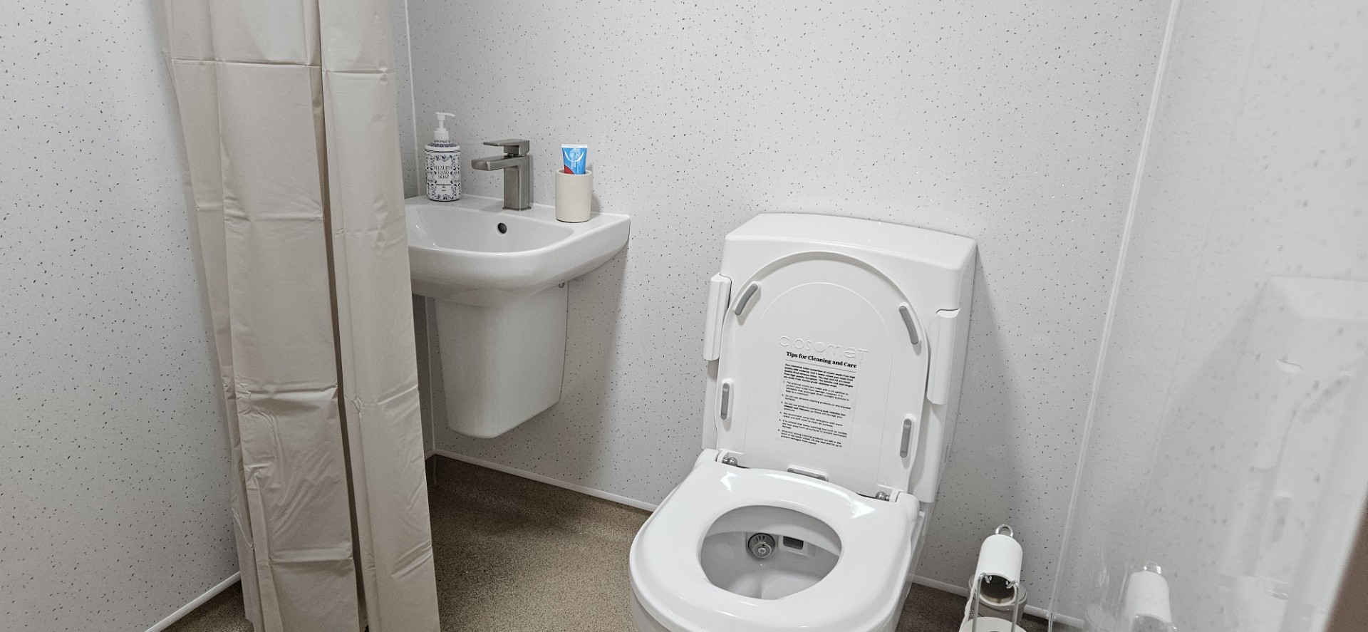 A clean, white bathroom with a toilet, a small sink, a shower curtain, soap dispenser, toilet paper, and a cup with toothbrushes on the sink. The overall space is simple and tidy.