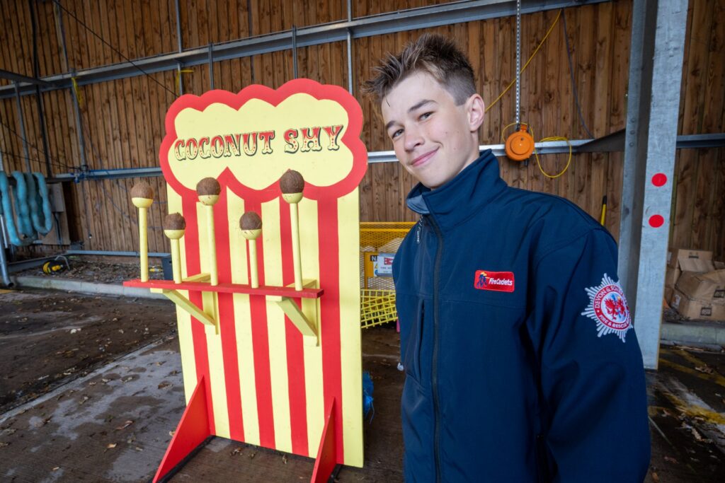 A smiling young person in a navy blue jacket stands beside a colorful "Coconut Shy" game with coconuts balanced on poles, inside a covered outdoor area with wooden walls.