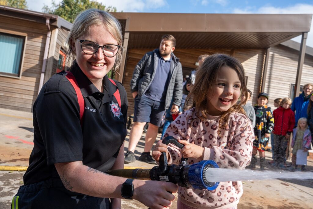 A smiling firefighter helps a young girl spray water from a hose, with a group of adults and children watching in the background outside wooden buildings on a sunny day.