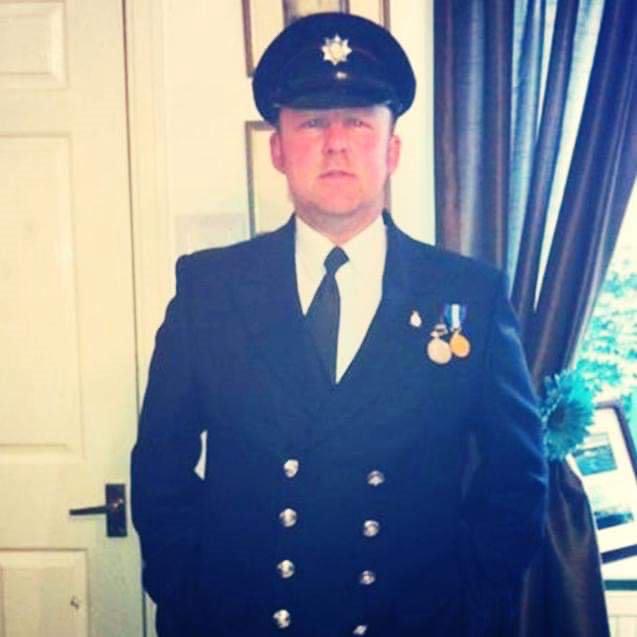 A person in a formal dark uniform and peaked cap stands indoors, facing the camera. The uniform has shiny buttons and medals pinned on the chest. There is a closed door and a window with curtains in the background.