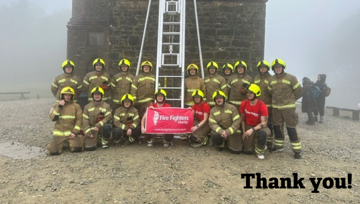 A group of firefighters in uniform and yellow helmets pose outdoors with a red Fire Fighters Charity banner in front of a stone building and tall ladder. 
