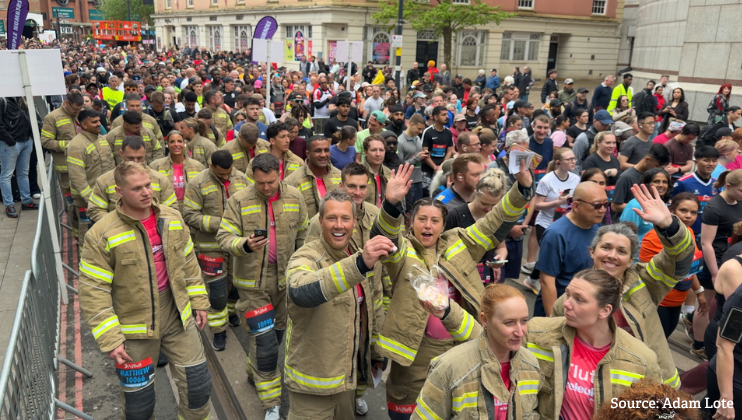 A group of firefighters in uniform walk among a large crowd of marathon runners on a city street. Some firefighters wave and smile at the camera. Spectators and participants fill the background.