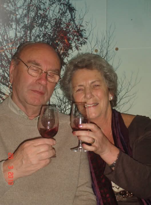 An older man and woman sit closely together, smiling and holding glasses of red wine. Leafless tree branches are visible in the background. The man wears glasses and a beige sweater; the woman wears a dark top and scarf.