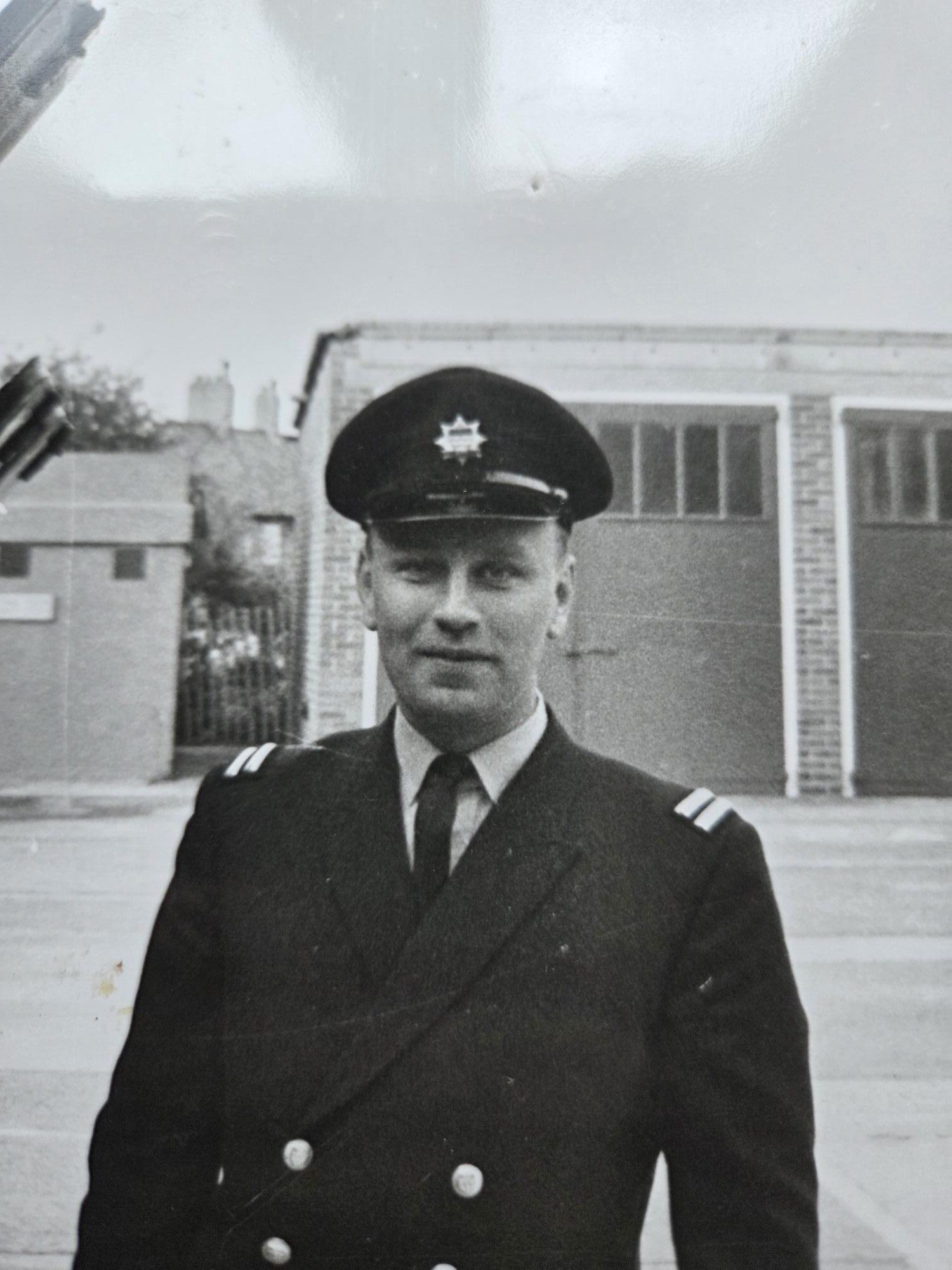 A man in a formal uniform with a peaked cap stands outside in front of brick buildings. The black and white photo has a vintage feel. The man looks toward the camera with a neutral expression.
