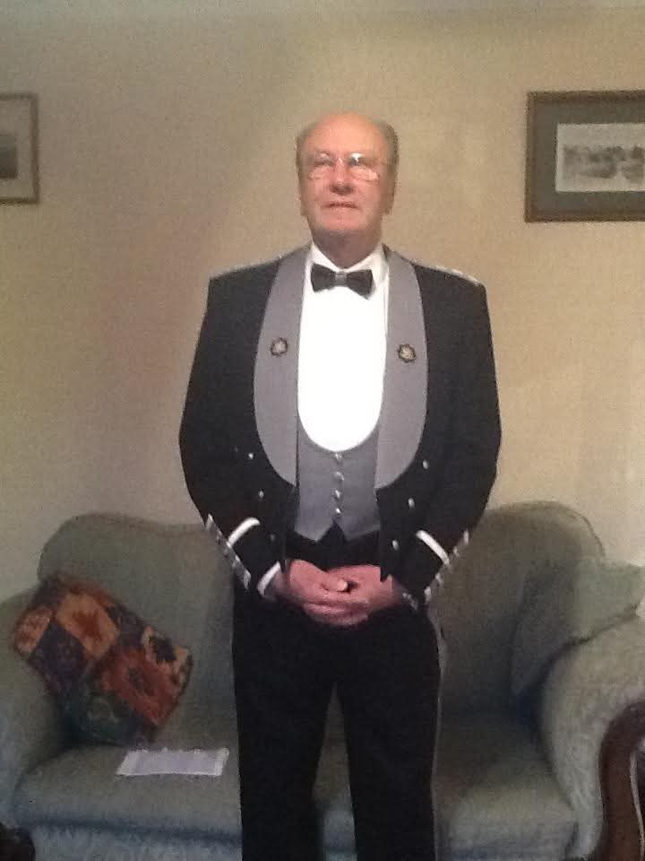 An older man stands in a living room wearing a formal black tuxedo with a bow tie, silver vest, and decorative buttons. He is standing in front of a sofa, with framed pictures on the wall behind him.