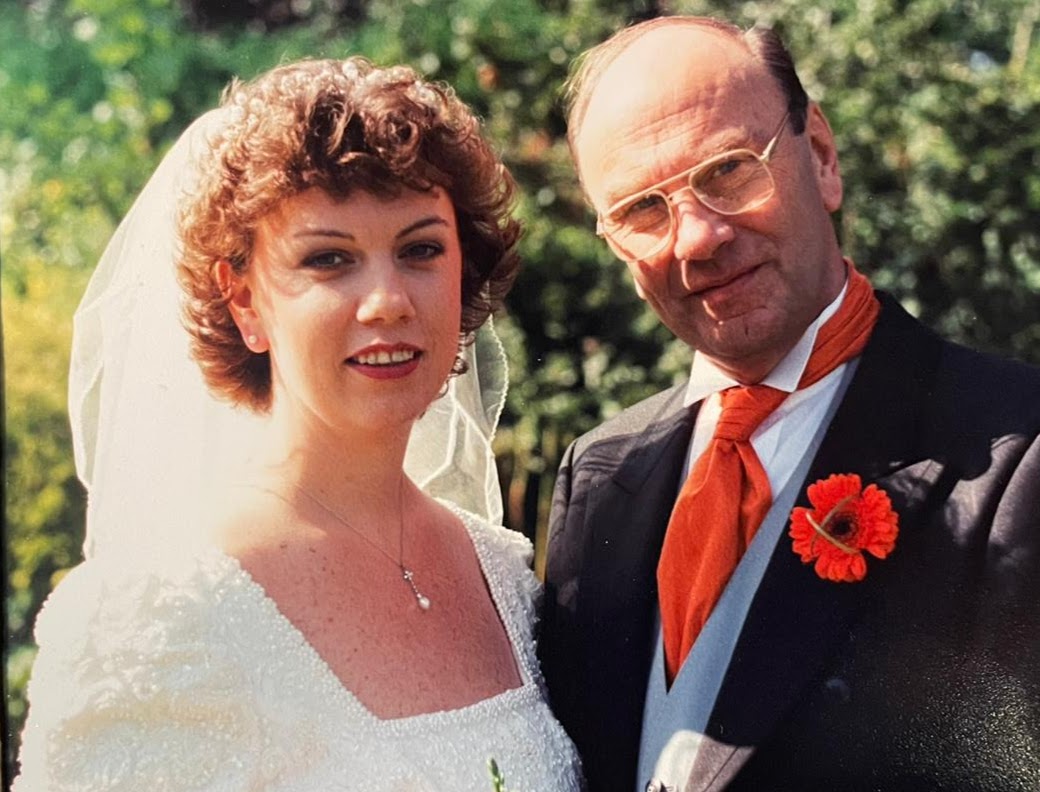 A woman in a white wedding dress and veil stands next to a man in a suit with an orange cravat and flower boutonniere, both smiling outdoors with greenery in the background.