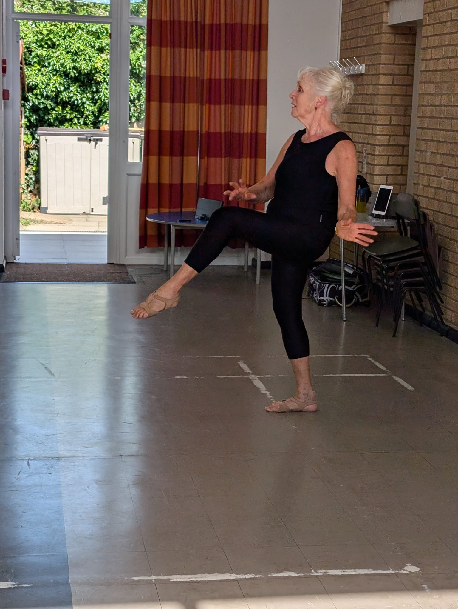 An older woman in black dancewear practices ballet indoors, standing on one leg with the other leg raised and arms outstretched. Sunlight streams in through an open door and window behind her.