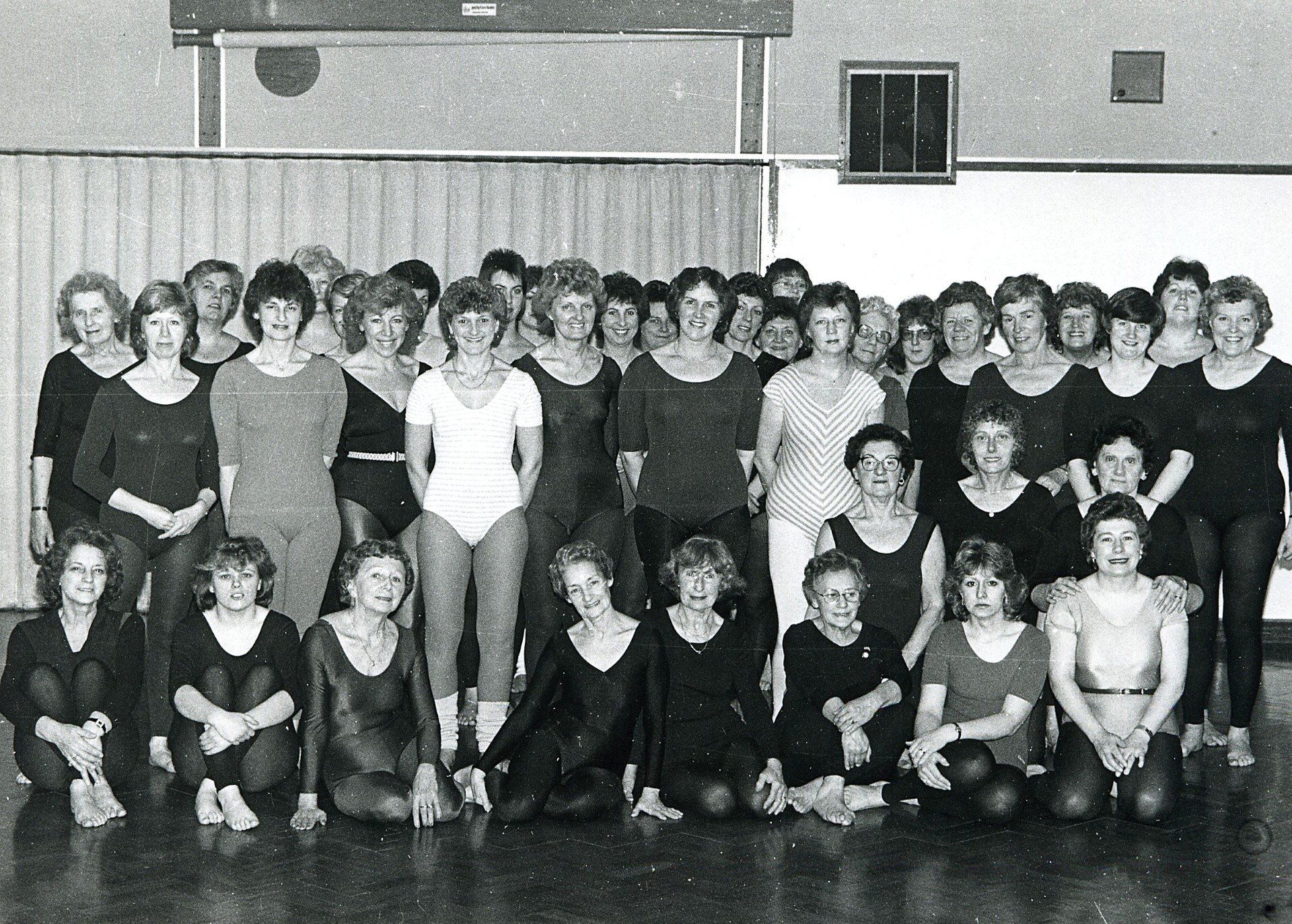 A black-and-white photo of around 30 women in leotards and workout attire posing together in a gymnasium, some standing and some sitting cross-legged on a wooden floor.