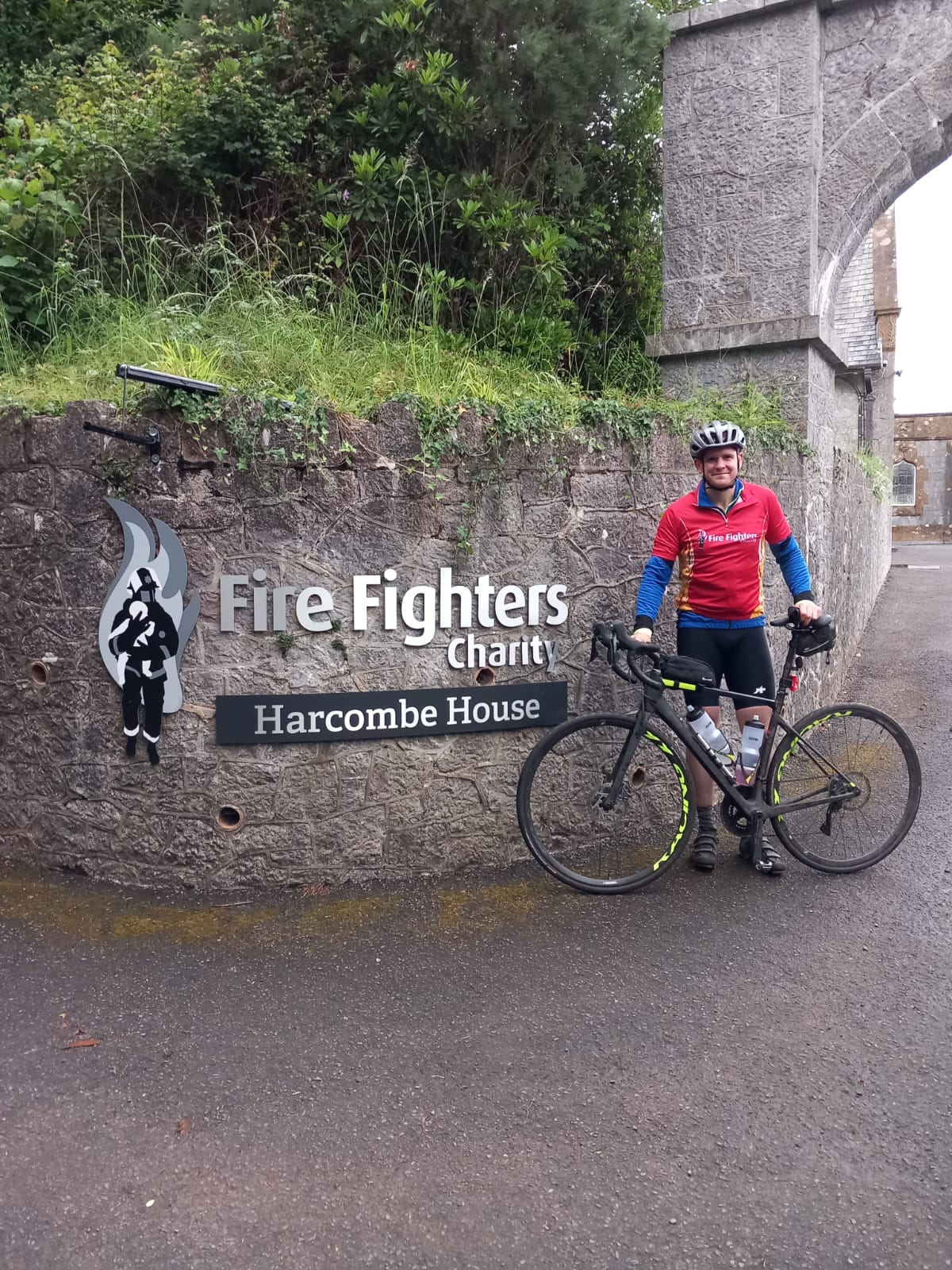 A cyclist in a red jersey and helmet stands with his bike in front of a stone wall with a "Fire Fighters Charity Harcombe House" sign. Trees and an arched stone entrance are visible in the background.