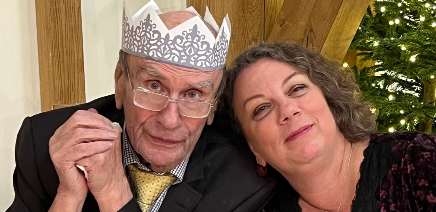 An elderly man wearing glasses, a suit, and a paper crown sits next to a smiling woman with curly hair. They are close together in front of a decorated Christmas tree.