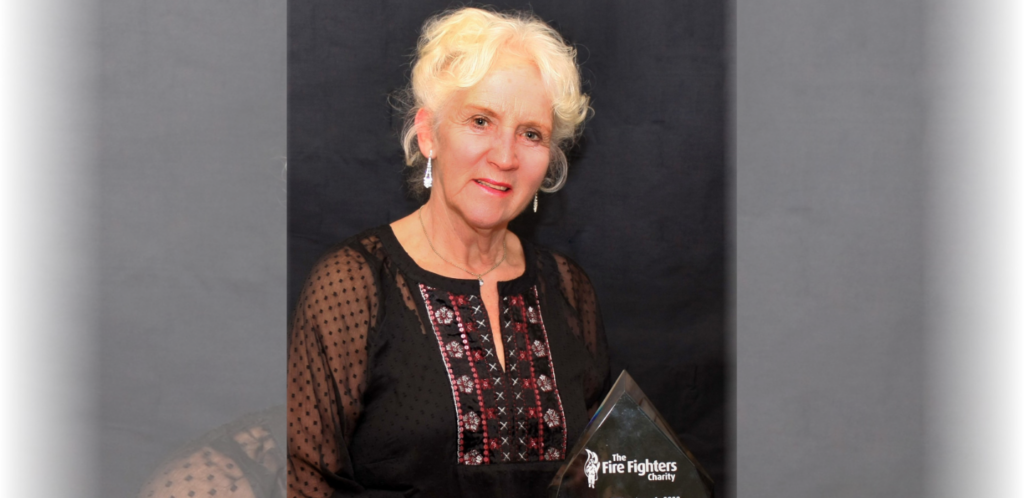 An older woman with curly white hair, wearing a black embroidered blouse, stands in front of a dark backdrop holding a glass award that reads 