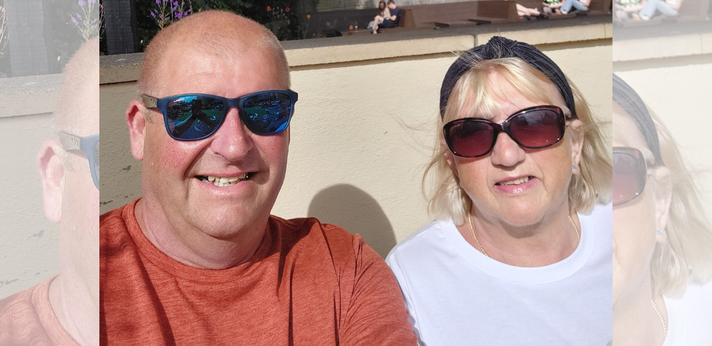 A smiling man in a red shirt and a woman in a white shirt, both wearing sunglasses, sit outside in bright sunlight with a beige wall behind them.