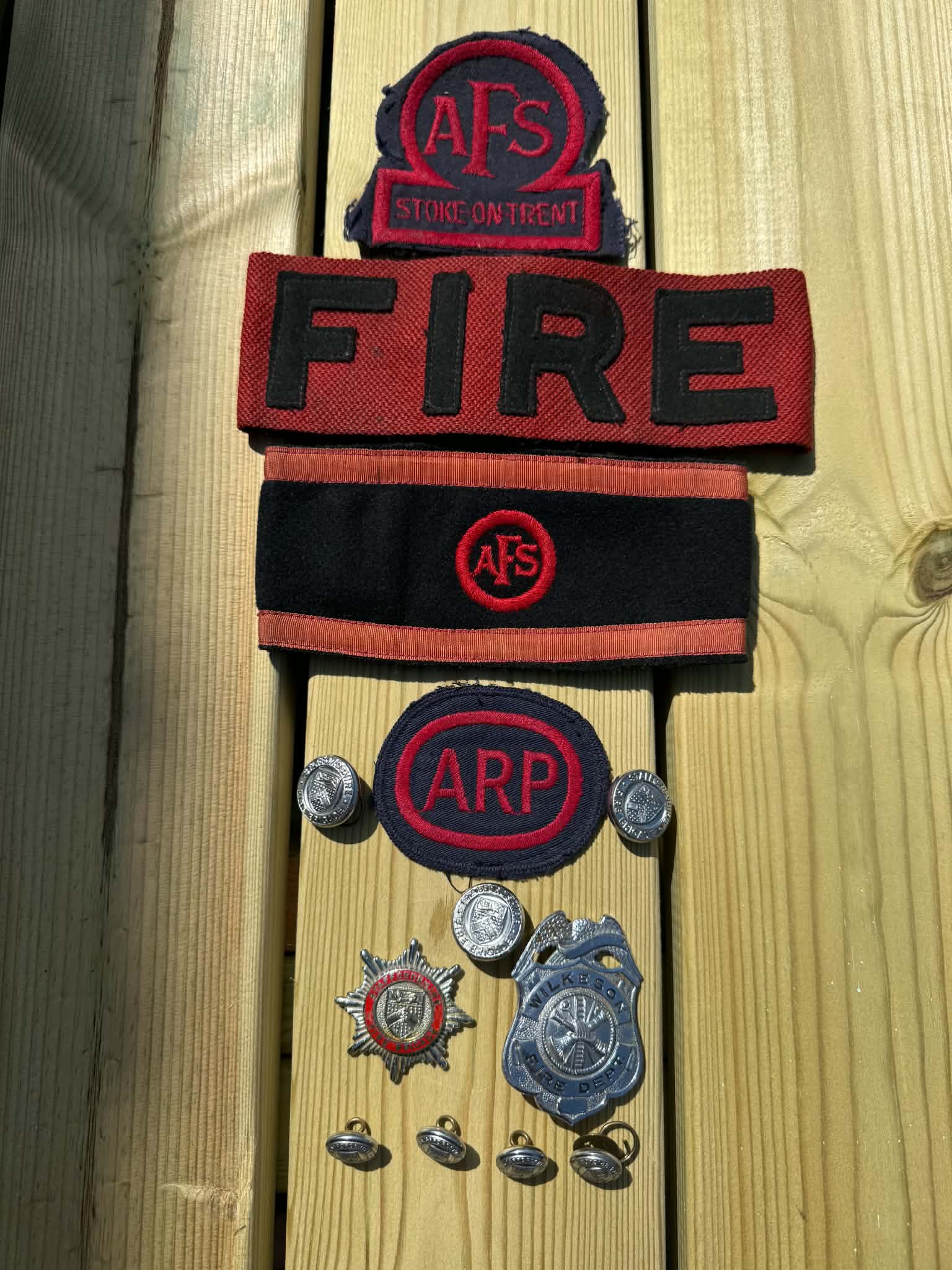 A collection of vintage fire service and civil defense badges, patches, and buttons, including “AFS” and “ARP” insignia, arranged neatly on a wooden surface.