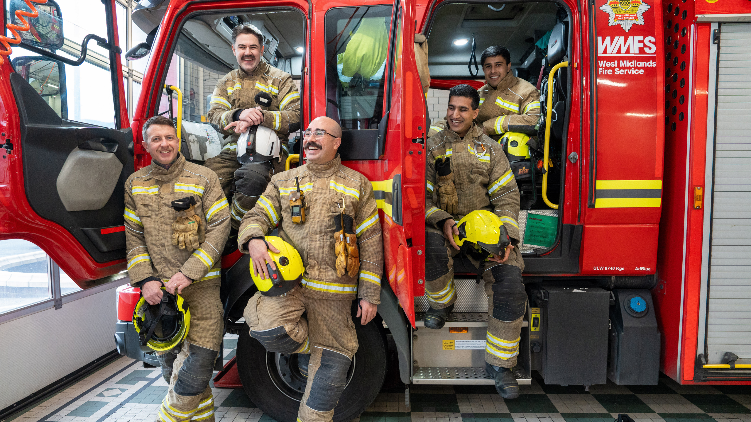 Five firefighters in uniform smile while standing and sitting on a bright red fire truck labeled 