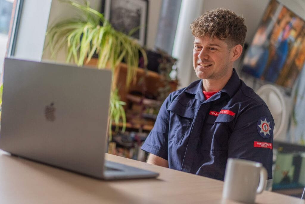A person in a fire and rescue uniform sits at a table, smiling while looking at a laptop. There are plants and shelves in the background in a well-lit room.