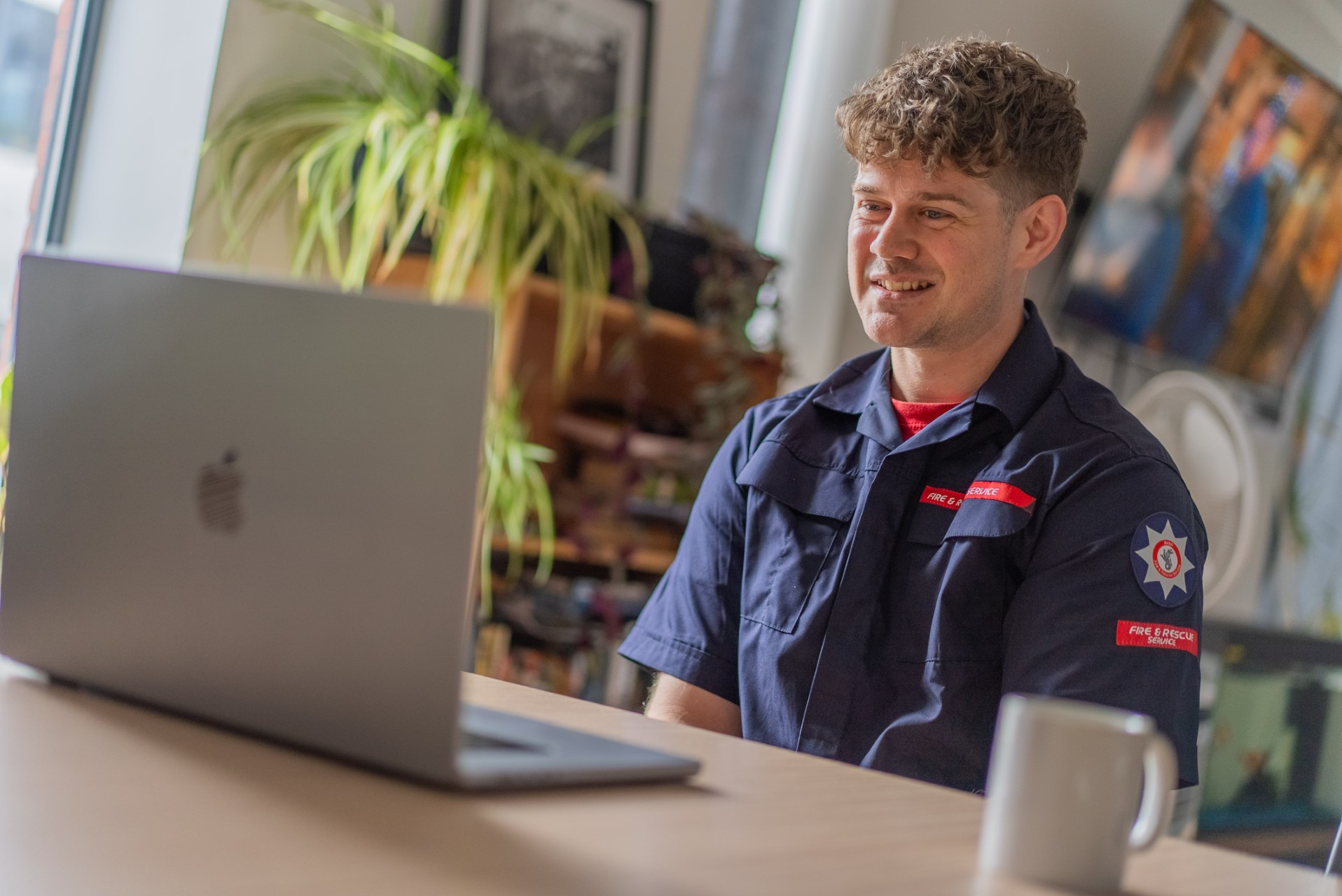 A person in a fire and rescue uniform sits at a table, smiling while looking at a laptop. There are plants and shelves in the background in a well-lit room.