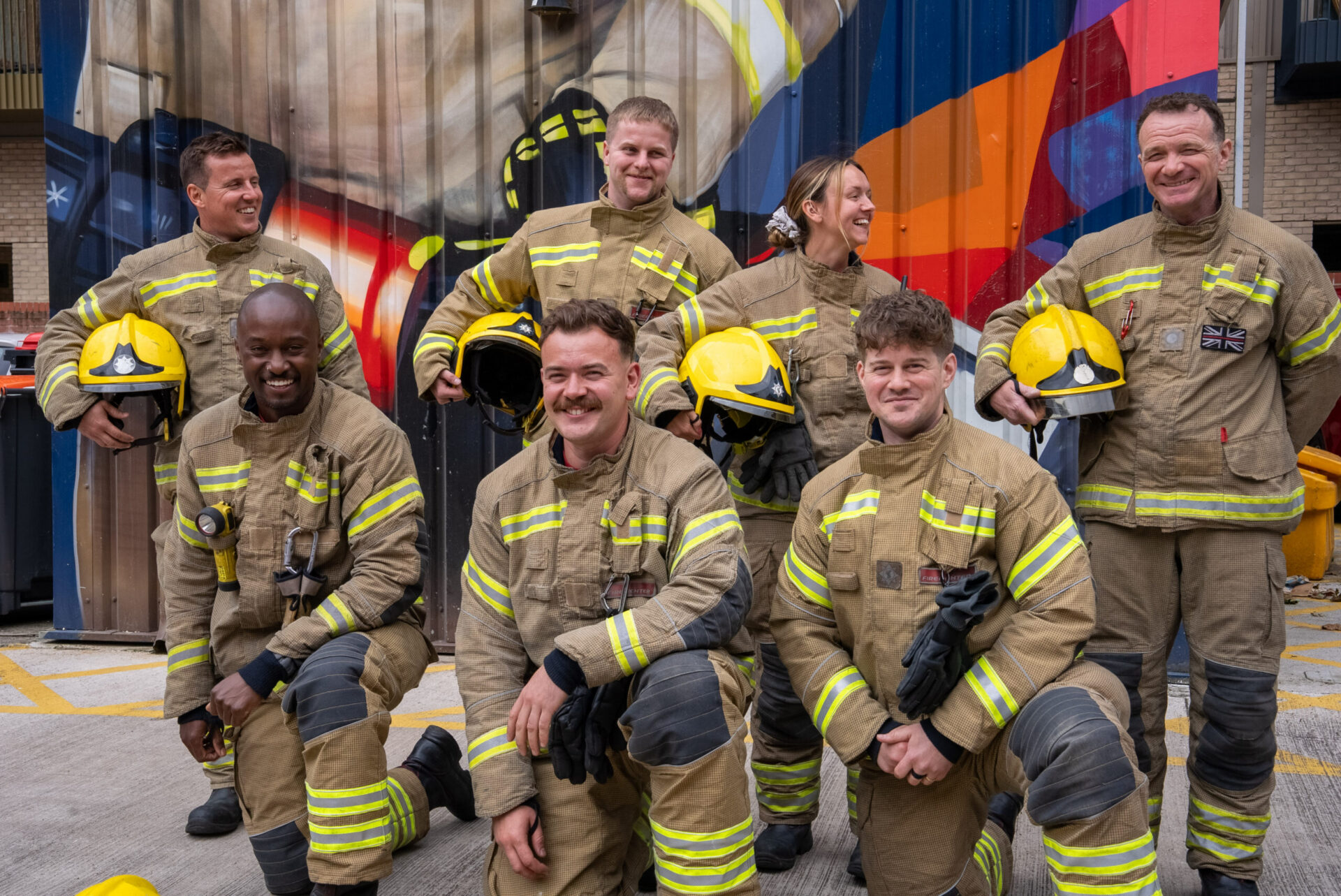Six firefighters in tan uniforms with yellow helmets pose and smile outside, in front of a colorful mural on a corrugated wall. Four are standing, and two are kneeling in the front row.