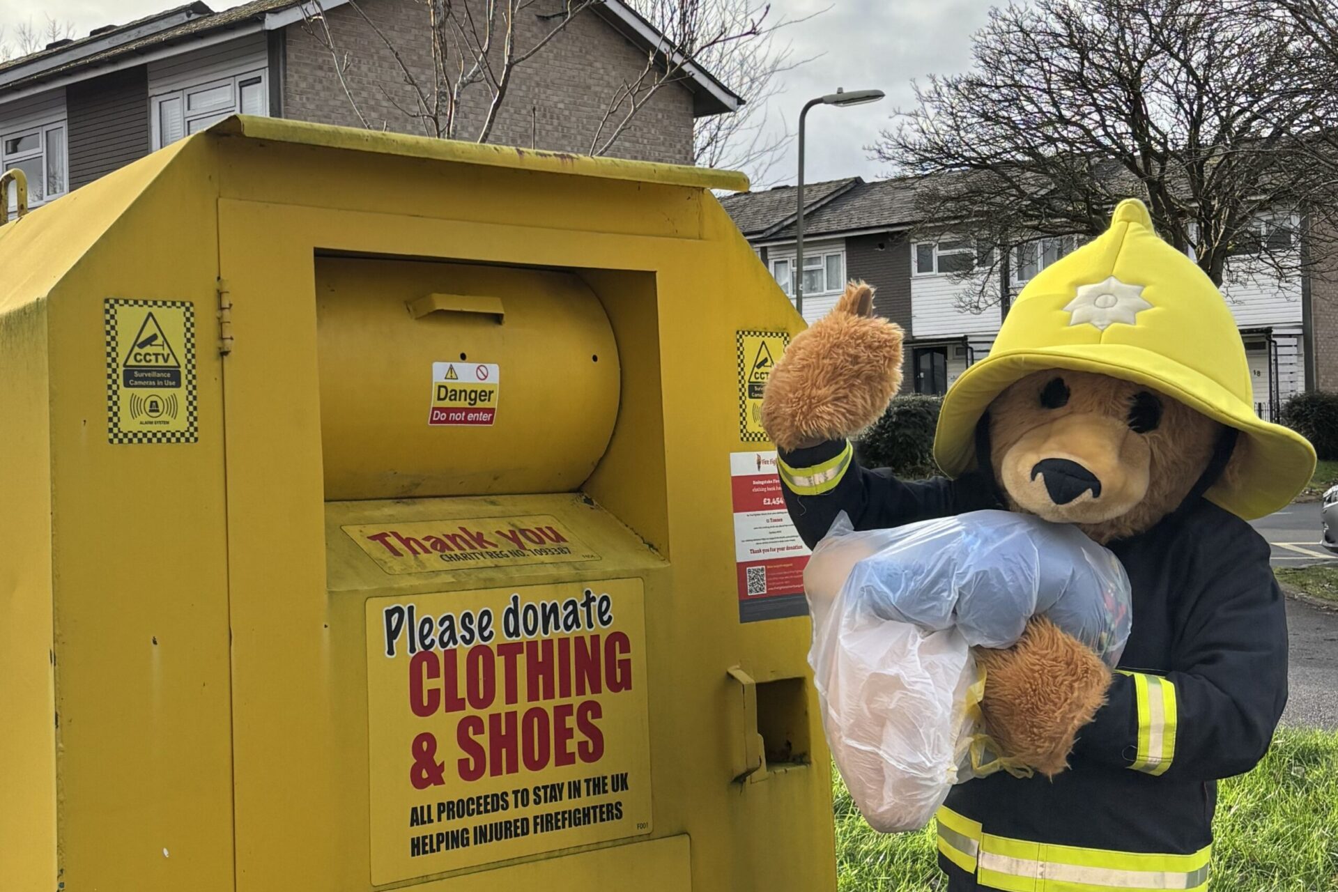 A person in a firefighter bear costume donates clothes at a yellow clothing and shoe donation bin on a suburban street, holding a clear bag of items and waving.