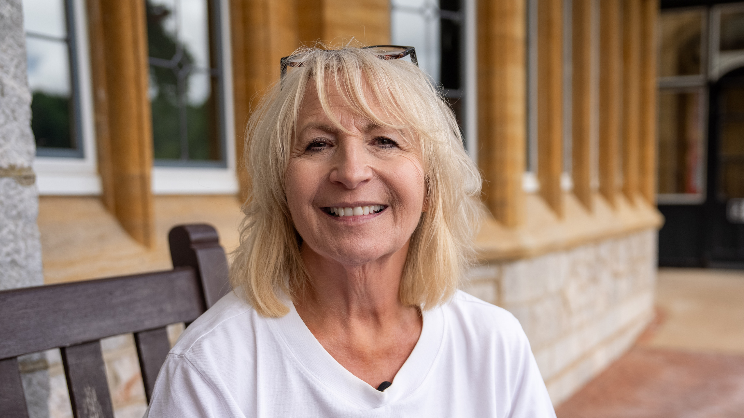 Smiling older woman with blonde hair sits on a wooden bench outside a Harcombe House with stone and yellow columns, wearing a white shirt and glasses on her head.