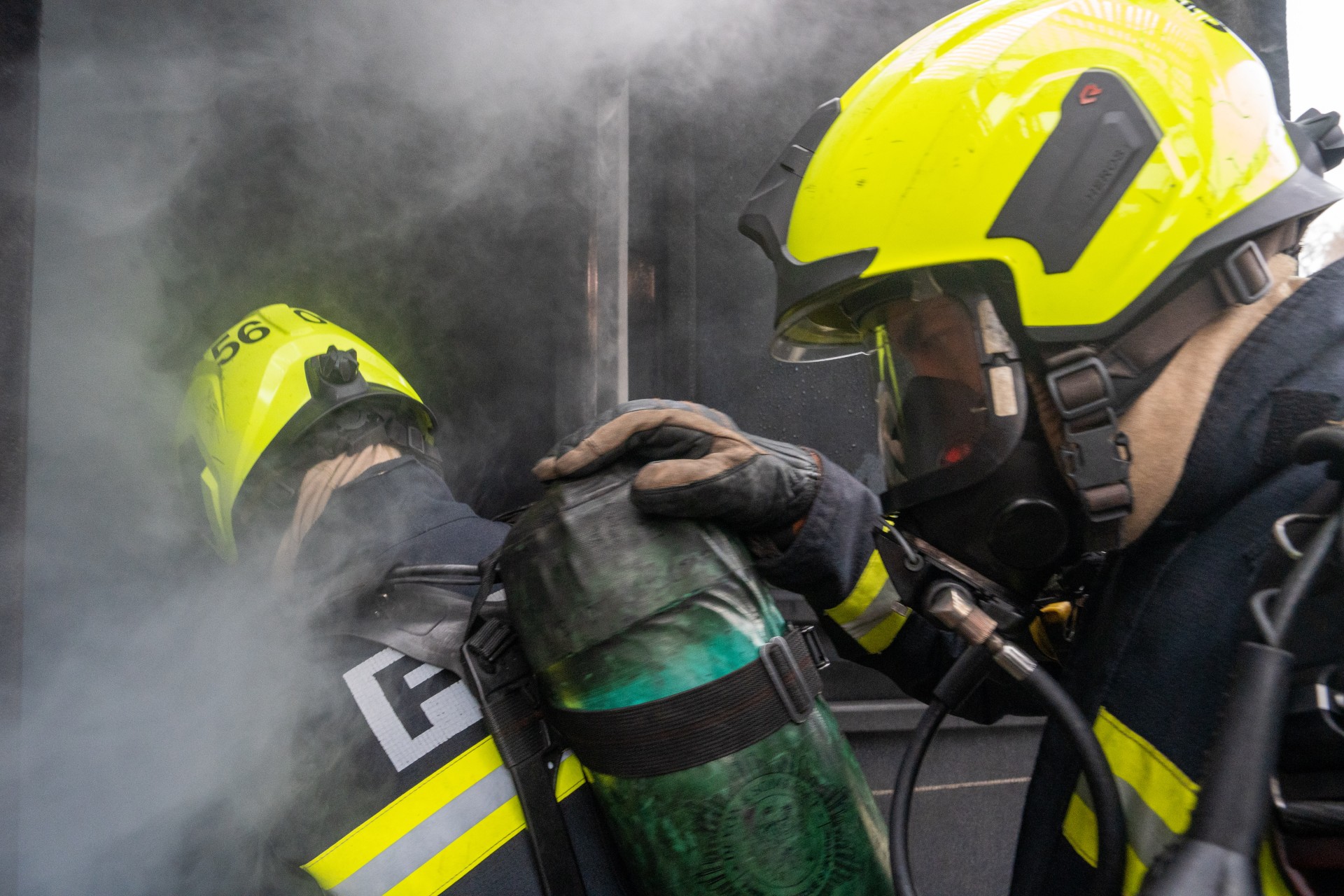 Two firefighters in yellow helmets and breathing gear enter a smoke-filled building, with one placing a hand on the other's shoulder for guidance and support.