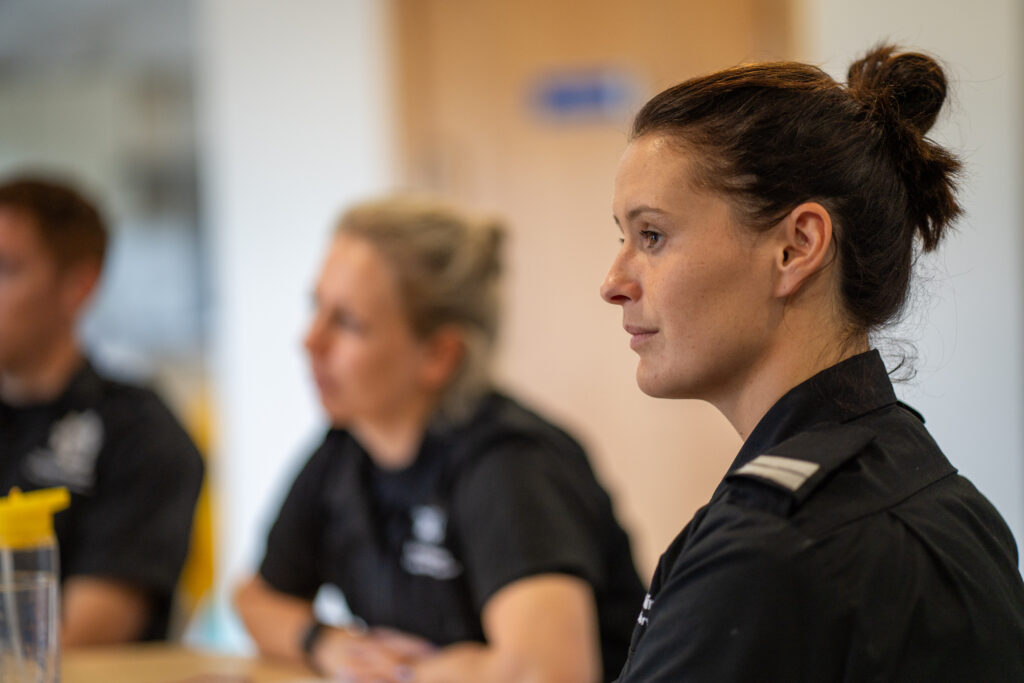 Three people in uniform sit at a table in a bright room, with a woman in focus looking attentively ahead. The background is blurred, highlighting her serious expression.