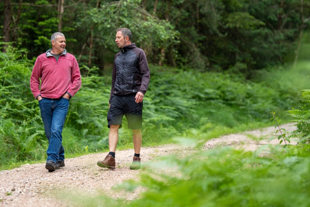 Two men walk and talk on a gravel path through a lush green forest. One wears a red sweater and jeans, the other a black jacket and shorts. They appear relaxed, enjoying nature and each other's company.