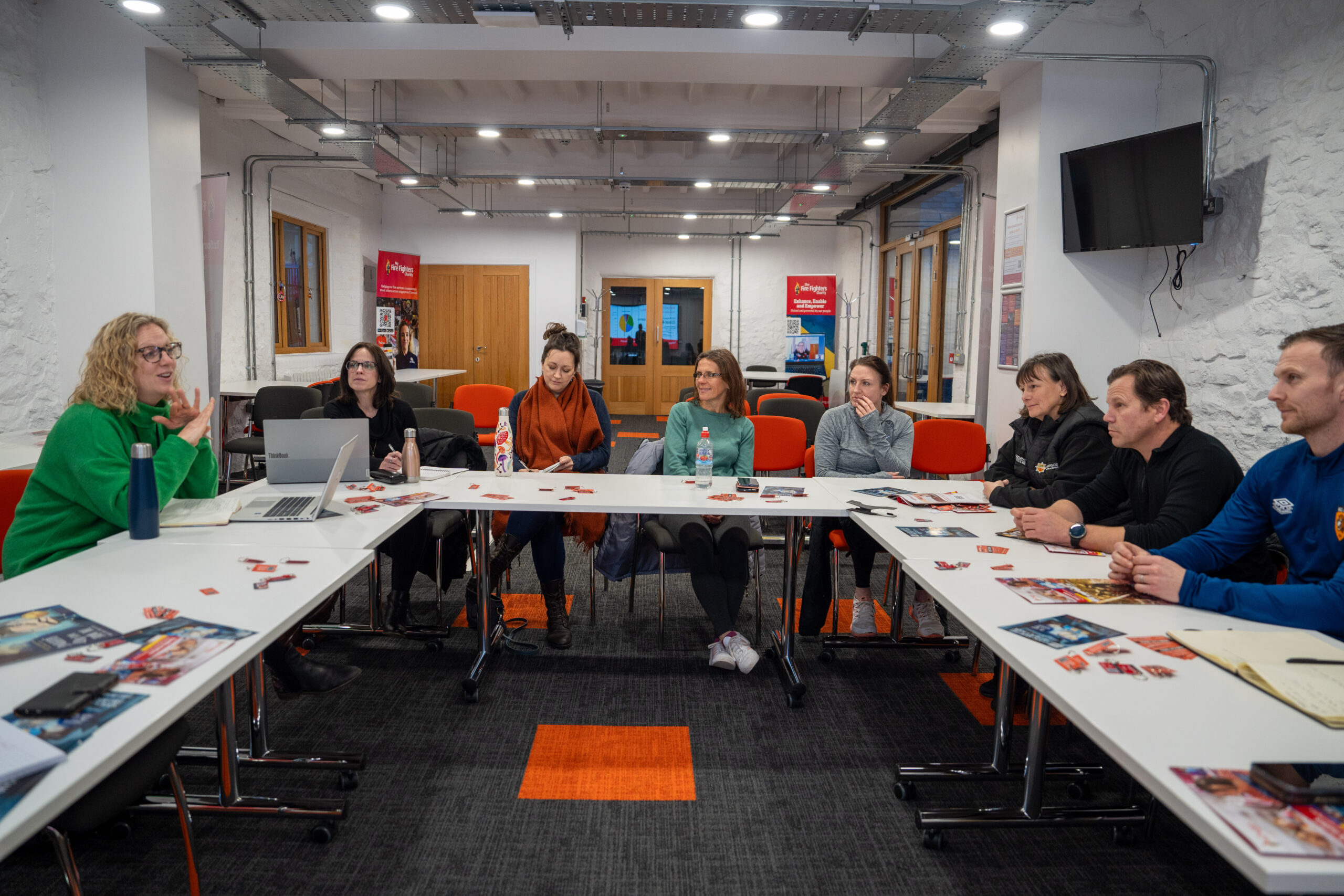 A group of people sit around tables in a meeting room, listening to a woman speaking. Papers and pamphlets are scattered on the tables. The room has white walls, red chairs, and visible ceiling pipes.