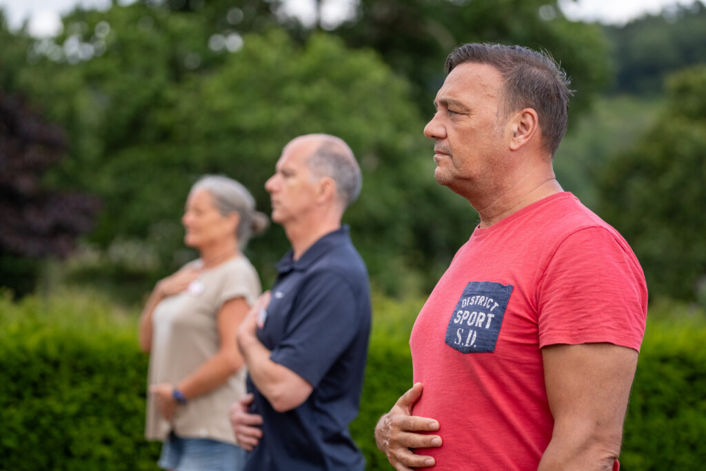 Three adults stand outdoors with eyes closed, one hand on their chest and one on their abdomen, appearing to practice mindfulness or deep breathing exercises, with greenery in the background.