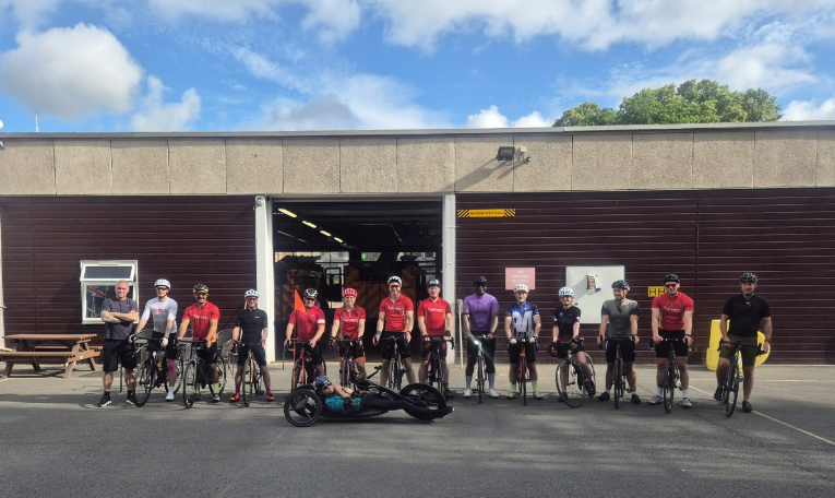 Twelve cyclists and one handcyclist pose in front of a large garage with brown doors on a sunny day; most wear helmets and cycling gear.