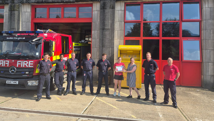 A group of firefighters and two civilians stand outside a fire station with a fire engine and a yellow box in the background. One civilian holds a certificate, and everyone is smiling at the camera.