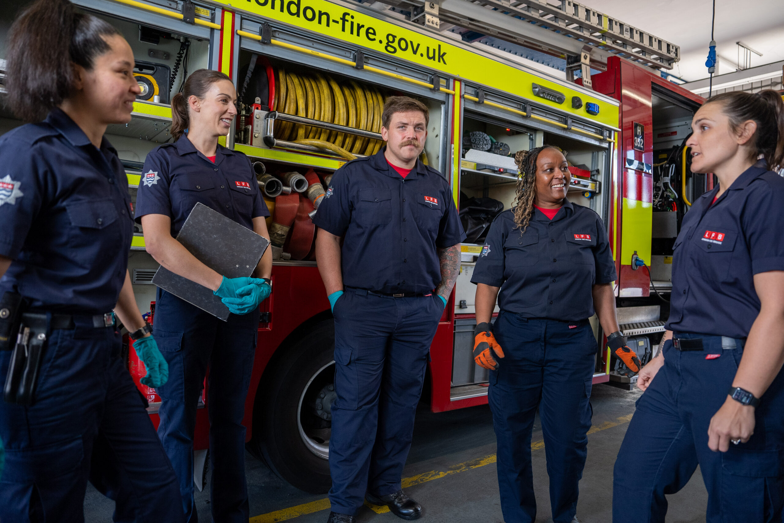 Five firefighters in uniform stand and talk near a fire engine, with tools and equipment visible on the truck. Four women and one man are smiling in a well-lit fire station.